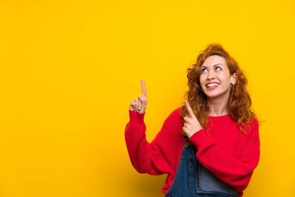 rothaarige Frau mit Overalls über isolierter gelber Wand, die mit dem Zeigefinger zeigt eine großartige Idee - Foto, Bild