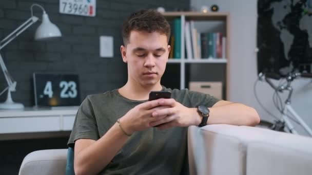 Stylish brunette caucasian teenager with a watch on his wrist is scrolling the feed in the app on his phone while sitting on a couch - Imágenes, Vídeo