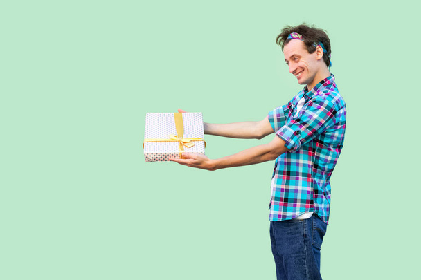 Side view of happy young hipster man in white t-shirt and checkered shirt holding present with yellow bow on green background - Foto, Imagem