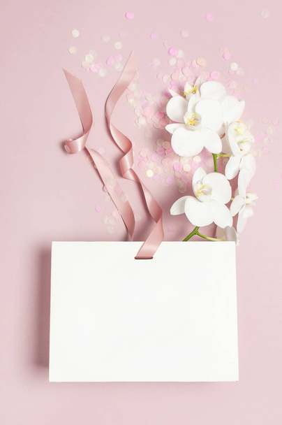Плоский вид сверху Белый фаленопсис орхидеи цветы праздничный конфетти белый подарочный мешок на розовом фоне. Открытка с орхидеей тропический цветок. Цветочная открытка на день рождения
 - Фото, изображение