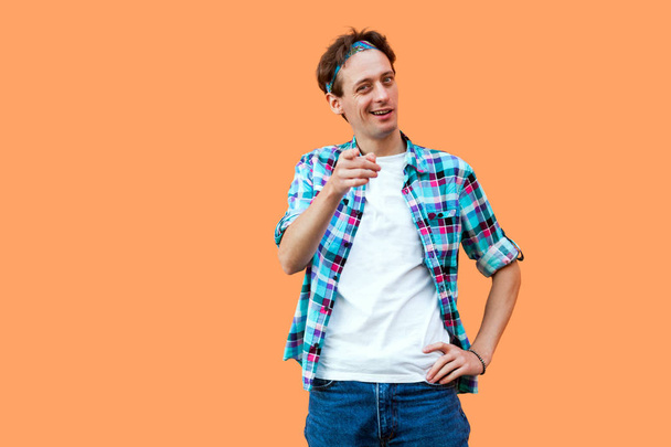 funny young man in casual blue checkered shirt and headband looking and pointing at camera with smile on orange background  - Zdjęcie, obraz