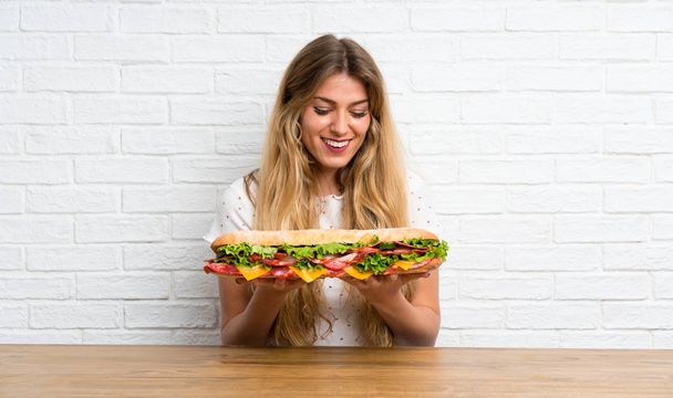 Gelukkige jonge blonde vrouw houdt een grote sandwich - Foto, afbeelding