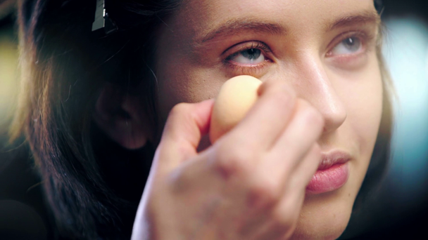cropped view of makeup artist applying concealer under eye on model face with cosmetic sponge - 映像、動画