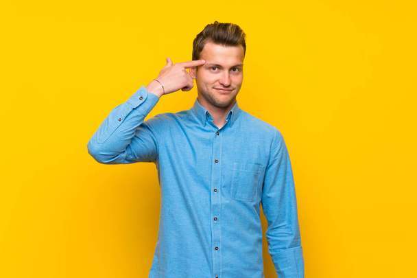 Hombre rubio sobre aislada pared amarilla haciendo el gesto de locura poniendo el dedo en la cabeza
 - Foto, Imagen