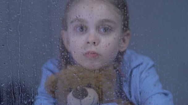 Abandonada menina pré-adolescente abraçando ursinho de pelúcia e olhando na janela no dia chuvoso
 - Filmagem, Vídeo