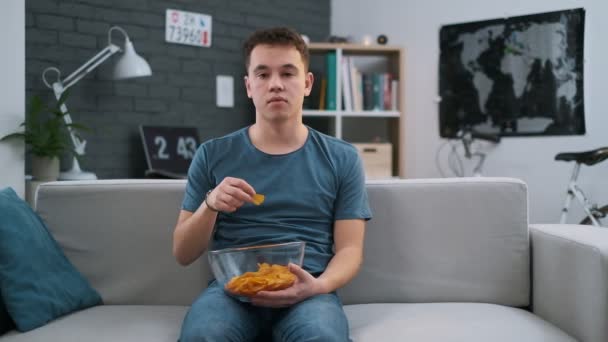 Front view of a bored teenager sitting on a sofa and eating chips at home - Materiał filmowy, wideo