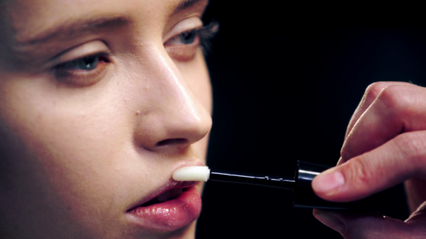 vista parcial del maquillador aplicando brillo labial brillante en los labios modelo con cepillo cosmético aislado en negro
 - Imágenes, Vídeo
