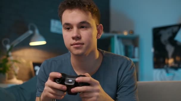 A hadsome teenager is sitting on a sofa and playing a video game on a playstation at home - Filmagem, Vídeo