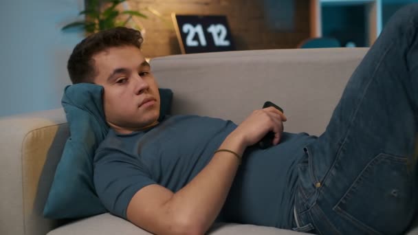A teenage boy is lying on the comfortable couch and watching TV, hes bored, so he switches channels with the remote control. - Кадри, відео