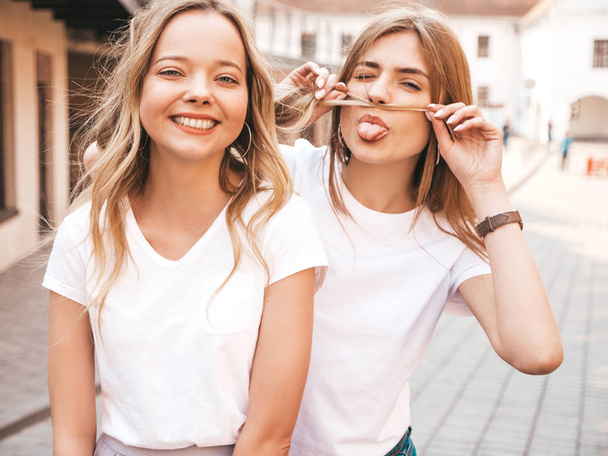 Portrait of two young beautiful blond smiling hipster girls in trendy summer white t-shirt clothes. Sexy carefree women posing in the street. Positive models having fun. Making mustache with hair - Zdjęcie, obraz