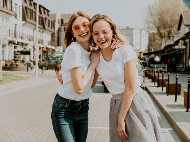 Portrait of two young beautiful blond smiling hipster girls in trendy summer white t-shirt clothes. Sexy carefree women posing on street background. Positive models having fun in sunglasses.Hugging - Fotografie, Obrázek