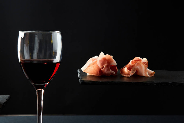 Composition of red wine in glass and pieces of meat snack on black background - Photo, image