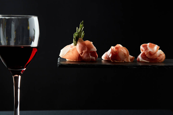 Composition of red wine in glass and pieces of meat snack on black background - Zdjęcie, obraz
