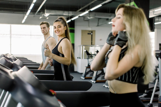 Young sports friends making cardio workout on stationary treadmill in the gym. Two girls and their handsome trainer running in machine treadmill at fitness gym club - Photo, image