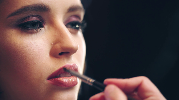 vista parcial del maquillador aplicando brillo de labios en labios modelo aislados en negro
 - Imágenes, Vídeo