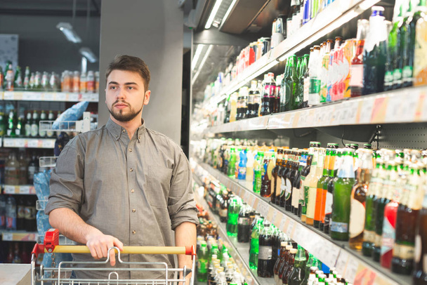 Muotokuva miehestä, jolla on kärry, seisoo supermarketin alkoholiosastolla lähellä hyllyjä olutpullojen kanssa. Oluen miesostaja supermarketista. Ostokset käsite
. - Valokuva, kuva