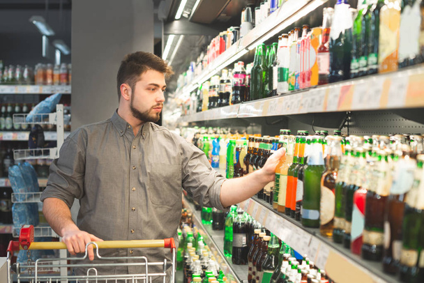 Un hombre con barba compra una cerveza en un supermercado, toma una botella de un estante en el departamento de alcohol. Hombre de negocios elige cerveza en un supermercado
. - Foto, imagen
