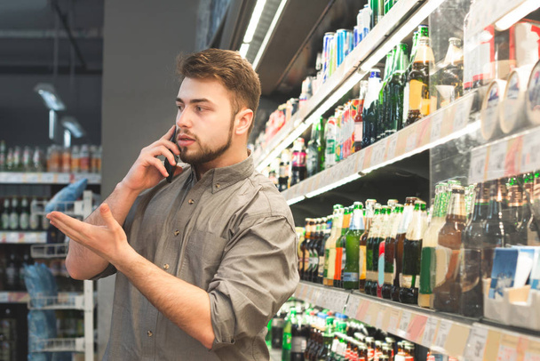 El hombre con su camisa está en los estantes con una cerveza en el supermercado y habla por teléfono. Hombre adulto discutiendo una compra en el supermercado por teléfono, selecciona el departamento de cerveza alcohólica
. - Foto, imagen