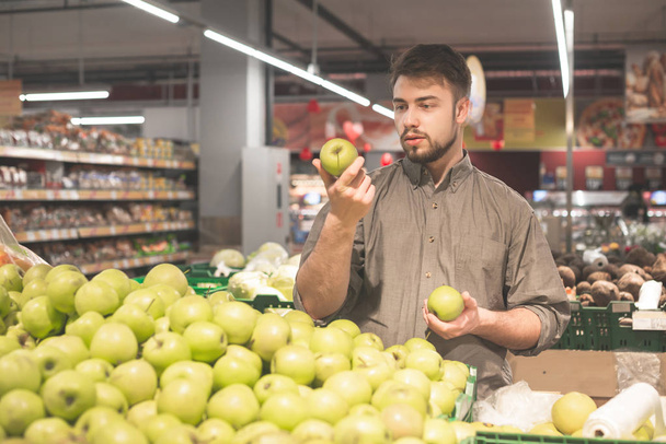 男はスーパーマーケットに立ち、リンゴを手に持ち、買い手はリンゴを買うシャツを着ている。あごひげを生やした男は、店内でリンゴを選び、手を見てリンゴを見る. - 写真・画像