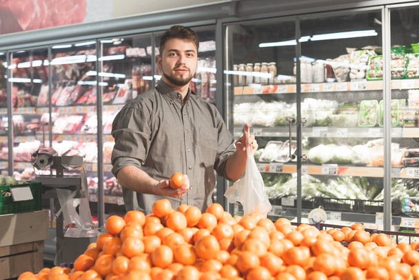 幸せな男は、スーパーマーケットでみかんを購入し、彼の手にパケットと柑橘類を持って立っています。笑顔のバイヤーはスーパーで果物を買います。スーパーマーケットを買い物し、商品を買う. - 写真・画像