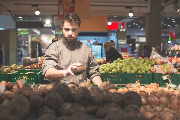 El hombre con manoplas selecciona verduras en un supermercado. Hombre adulto cavando ingredientes para platos en una tienda de comestibles, un departamento de verduras. Comprador compra verduras
. - Foto, Imagen