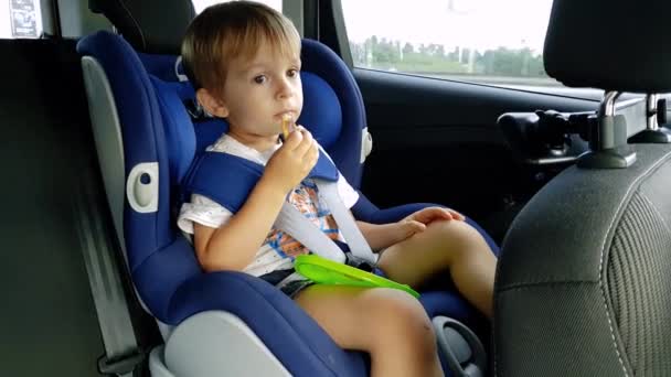 4k video of little toddler boy eating while riding in car. Child sitting in safety car seat and eating cookie - Footage, Video
