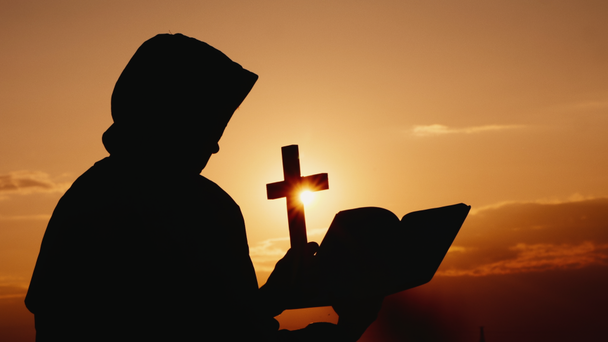 Ένας μοναχός σε μια κουκούλα με ένα σταυρό στα χέρια του στέκεται στο φόντο ενός δραματικού ουρανού στο ηλιοβασίλεμα - Πλάνα, βίντεο