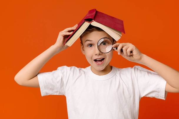 Χαριτωμένο έξυπνο αγόρι με μεγεθυντικό φακό στο χέρι κρύβονται κάτω από το βιβλίο - Φωτογραφία, εικόνα