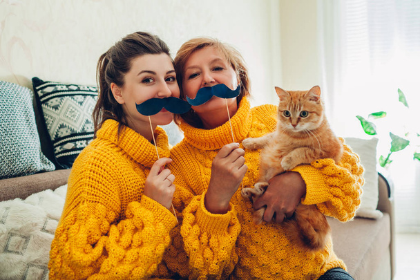 Vanhempi äiti ja hänen aikuinen tyttärensä ottavat selfien kissan kanssa käyttäen valokuvauskopin rekvisiittaa kotona. Äitienpäivän käsite
. - Valokuva, kuva