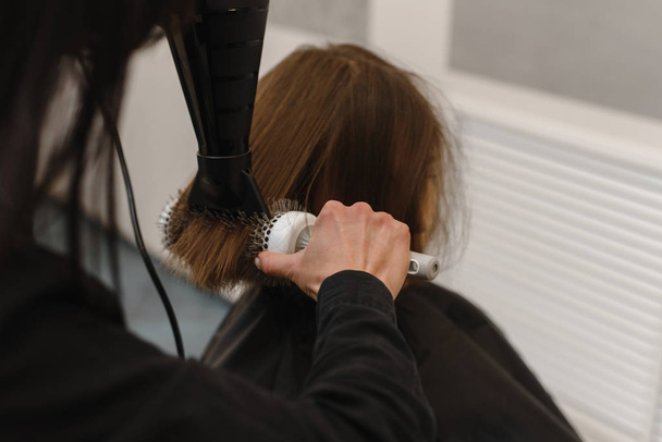 το κομμωτήριο στεγνώνει τα βρεγμένα μαλλιά με ένα πιστολάκι για τα μαλλιά στον πελάτη. Όμορφο σαλόνι ομορφιάς. Χώρος αντιγραφής - Φωτογραφία, εικόνα