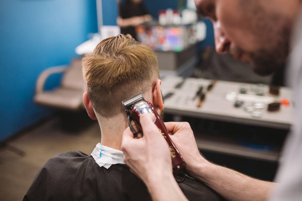 Tiro de perto de um homem a cortar o cabelo na barbearia. Cabeleireiro masculino servindo cliente, fazendo corte de cabelo usando máquina e pente
 - Foto, Imagem