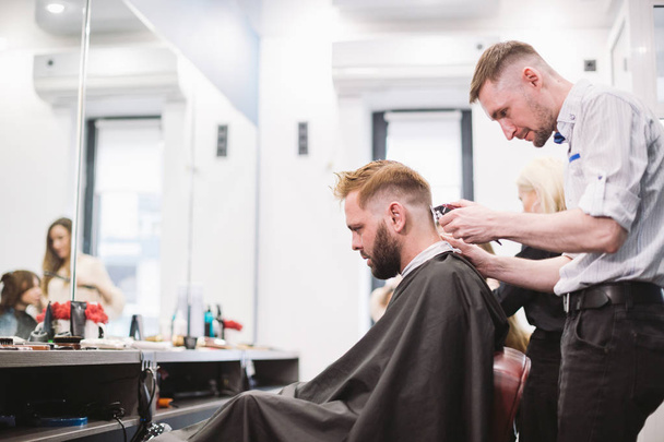 Крупный план человека, стригущегося в парикмахерской. Мужчина парикмахер, обслуживающий клиента, делает стрижку с помощью машины и расчески
 - Фото, изображение