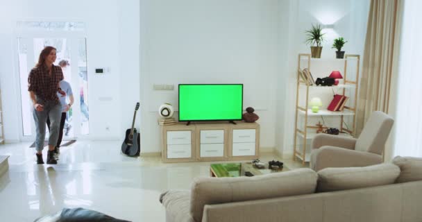 Charismatic group of friends coming from outside in the house sitting quickly on the sofa and switch on the TV to watching a spot match , green screen on the TV - Кадры, видео