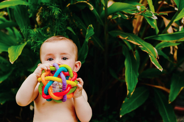 Baby sitting on the barefoot grass nibbling on a plastic toy. - 写真・画像