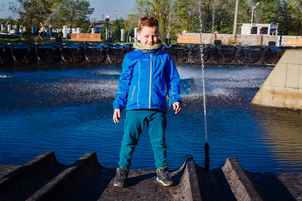 seven-year-old boy in a blue windbreaker stands near the city fountain in the spring - Fotoğraf, Görsel