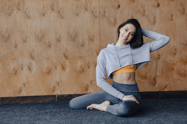 Menina fitness atraente jovem sentado no chão perto da janela no fundo de uma parede de madeira, descansando em aulas de ioga
 - Foto, Imagem