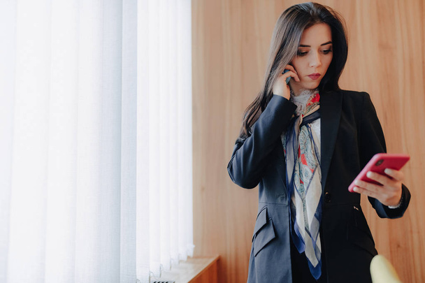 Młoda dziewczyna emocjonalna atrakcyjna w stylu biznesowym ubrania w oknie z telefonem w nowoczesnym biurze lub audytorium - Zdjęcie, obraz
