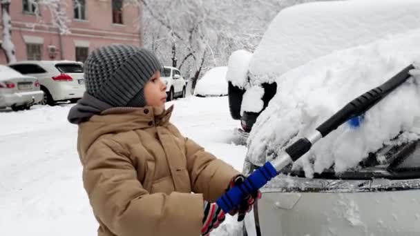 wideo 4K malucha chłopiec czyszczenia samochodu z śniegu z pędzlem dziecko pomaga czyszczenia pojazdu w godzinach porannych po zamieć - Materiał filmowy, wideo