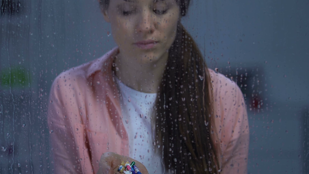 Lonely sick woman holding palm of medication near rainy window looking to camera - Video
