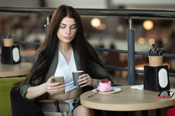 Retrato de una mujer feliz comiendo un pedazo de pastel y tomando café mientras está sentada a la mesa en un café adentro - Foto, Imagen