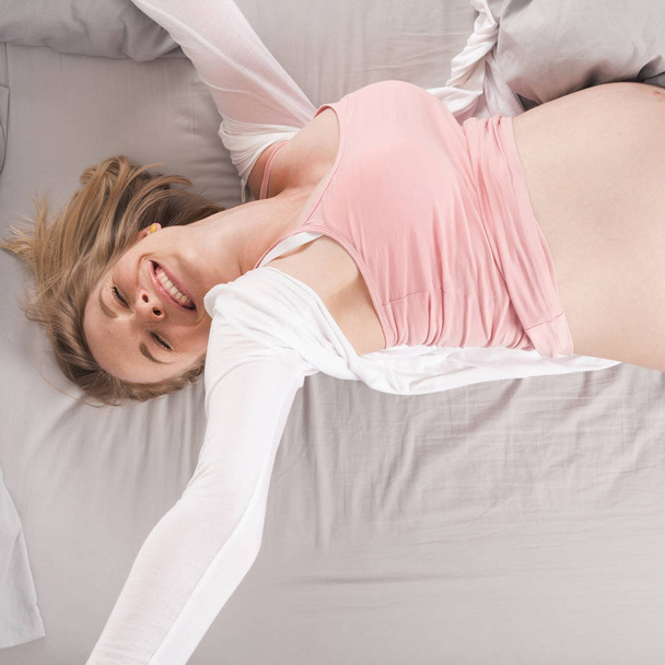 Ευτυχισμένος νεαρή έγκυο γυναίκα βρίσκεται στο κρεβάτι και το τέντωμα. Το Top view - Φωτογραφία, εικόνα