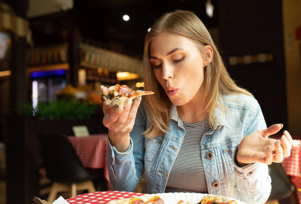 Funny blonde girl in jeans jacket eating pizza at restaurant. - Фото, изображение