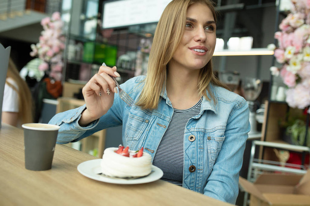 Прекрасная улыбающаяся молодая женщина ест торт и пьет кофе в кафетерии
.  - Фото, изображение