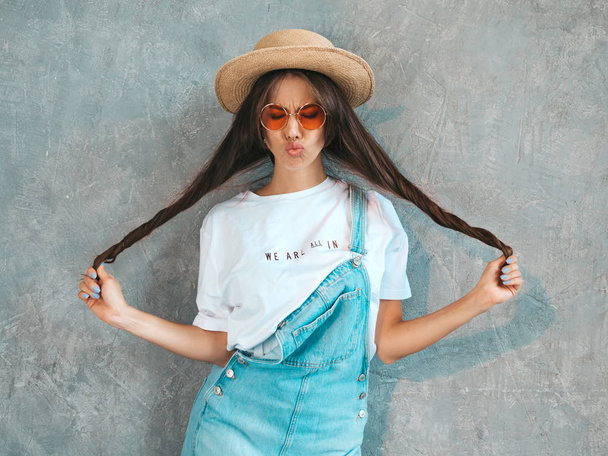 Young beautiful smiling woman looking at camera. Trendy girl in casual summer overalls clothes and hat. Funny and positive female posing near gray wall in studio. Playing with her hair - Foto, Imagem
