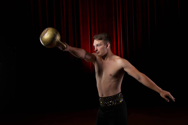 Power gymnast circus with weight.Handsome guy with a muscular body holding a kettlebell on a black stage background - Photo, Image