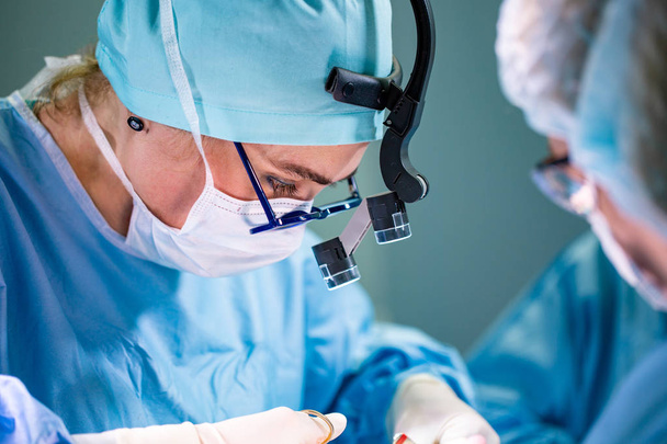 Chirurg und seine Assistentin bei der Schönheitschirurgie im Operationssaal des Krankenhauses. Chirurg mit Maske trägt Lupen während medizinischer Prozedur. - Foto, Bild