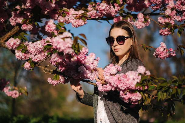 Gyönyörű lány blézer és napszemüveg állni a Sakura fa. Gyönyörű rózsaszín fa a parkban - Fotó, kép