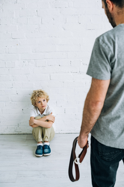 cropped view of father holding belt and sad son sitting on floor - Photo, Image