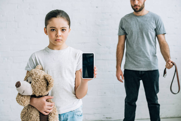 partial view of abusinve father and daughter with teddy bear showing smartphone with blank screen - Photo, Image