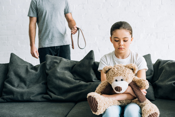 partial view of abusive father with belt and sad daughter with teddy bear sitting on sofa - Foto, Imagem